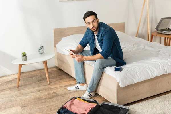 Viajero joven con tableta digital mirando a la cámara cerca de la maleta y el pasaporte en el dormitorio - foto de stock