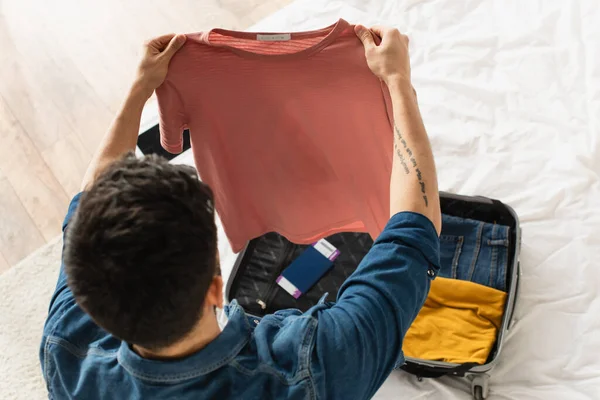 Visão aérea do homem segurando t-shirt perto da mala com passaporte na cama — Fotografia de Stock