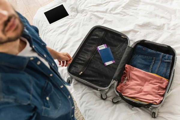 Vista de alto ângulo do homem desfocado em pé perto da mala com passaporte e tablet digital na cama — Fotografia de Stock