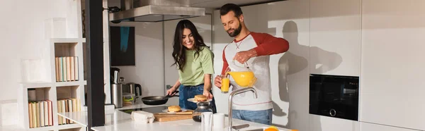 Man pouring orange juice near smiling girlfriend with pancakes in kitchen, banner — Stock Photo
