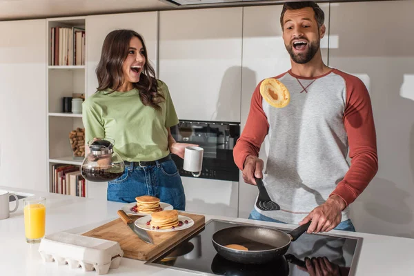 Excited man cooking pancakes near girlfriend with coffee in kitchen — Stock Photo