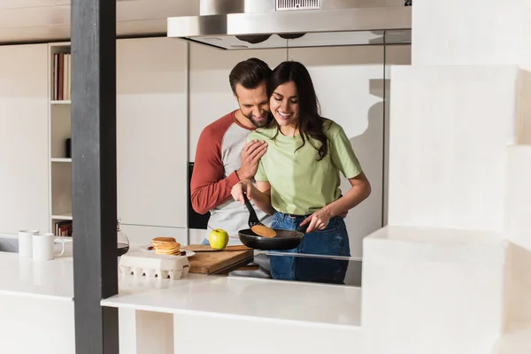 Усміхнений чоловік обіймає дівчину готуючи млинці на кухні — стокове фото