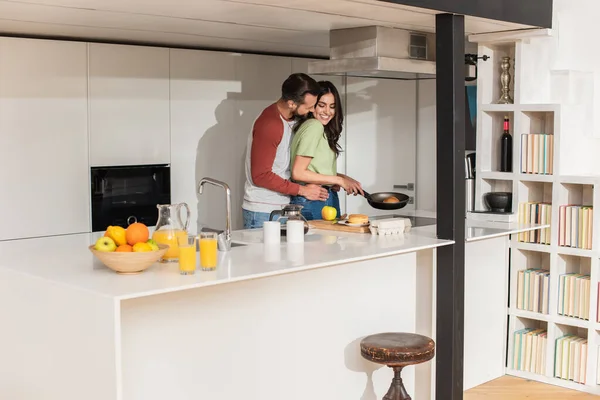 Homem abraçando namorada cozinhar panquecas perto de frutas e bebidas na cozinha — Fotografia de Stock