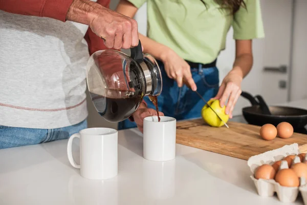 Ausgeschnittene Ansicht eines Mannes, der Kaffee in der Nähe verschwommener Freundin einschenkt, die in Küche Apfel schneidet — Stockfoto