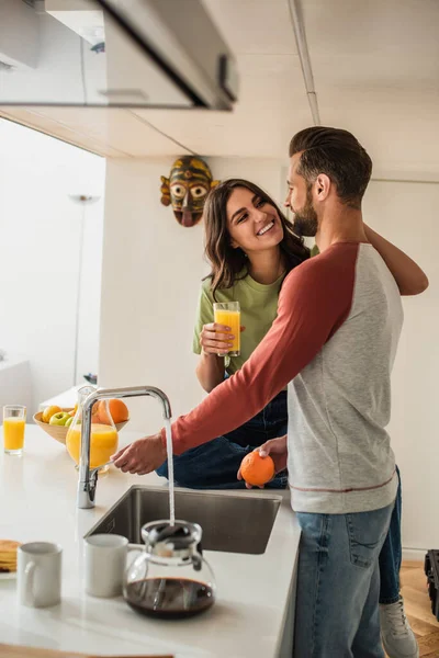 Smiling woman with orange juice looking at boyfriend with orange in kitchen — Stock Photo