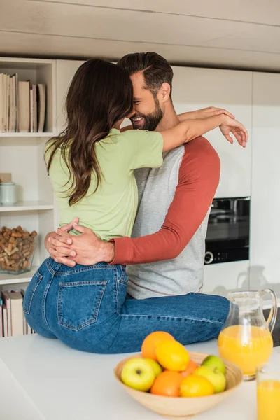 Uomo sorridente che abbraccia la ragazza vicino al succo d'arancia e frutta sfocata in cucina — Foto stock