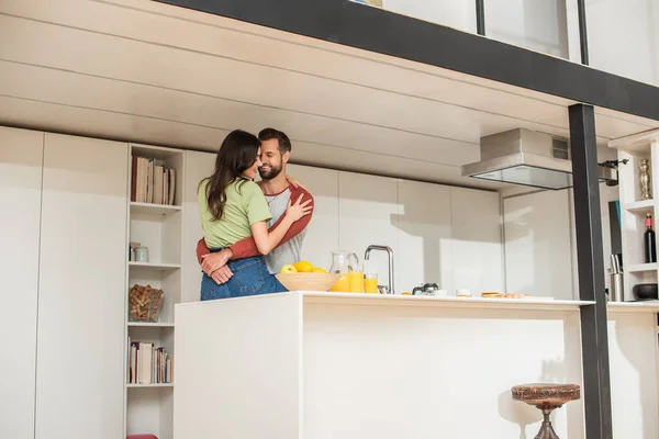 Sorrindo homem abraçando namorada perto do café da manhã com suco de laranja na cozinha — Fotografia de Stock