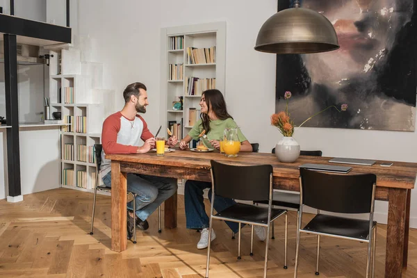 Vista lateral de una pareja sonriente sentada cerca del desayuno y computadoras portátiles en la mesa - foto de stock