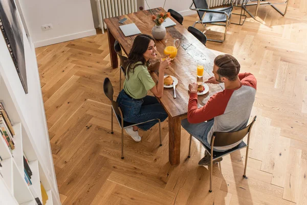 Вид сверху на улыбающуюся женщину с апельсиновым соком, сидящую рядом с парнем во время завтрака дома — стоковое фото