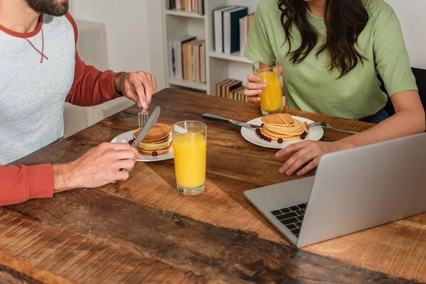 Vista ritagliata della coppia seduta vicino al succo d'arancia, frittelle e laptop sul tavolo — Foto stock