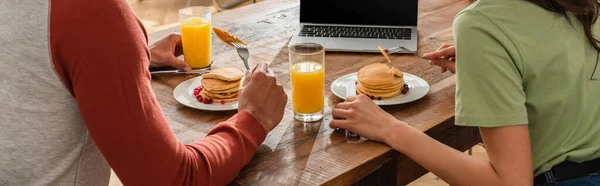 Ausgeschnittene Ansicht eines Paares beim Frühstück mit Pfannkuchen in der Nähe von Laptop, Banner — Stockfoto