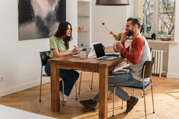 Man with coffee talking on smartphone near girlfriend writing on notebook and laptops — Stock Photo