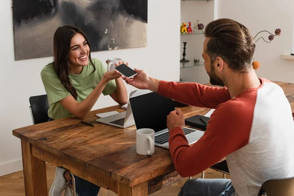 Hombre dando teléfono inteligente a la novia sonriente con café cerca de las computadoras portátiles en casa - foto de stock