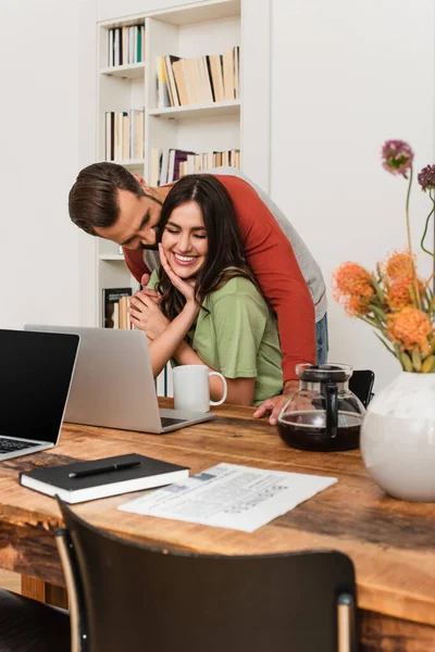 Hombre abrazando novia sonriente cerca de computadoras portátiles, café y periódico en casa - foto de stock