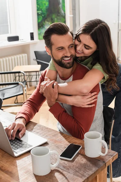Mujer alegre abrazando novio usando el ordenador portátil cerca de teléfono inteligente y tazas - foto de stock