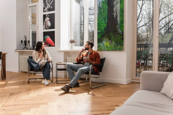 Smiling woman with book looking at boyfriend with smartphone — Stock Photo