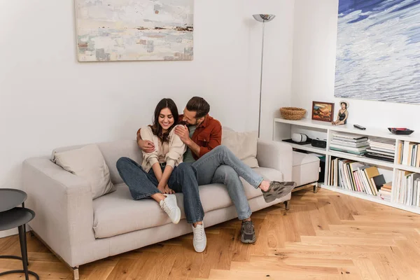 Sonriente hombre abrazando novia en el sofá en la sala de estar - foto de stock