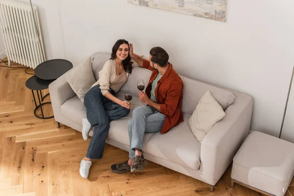 Vista aérea del hombre con vino ajustando el pelo de la novia sonriente en el sofá - foto de stock