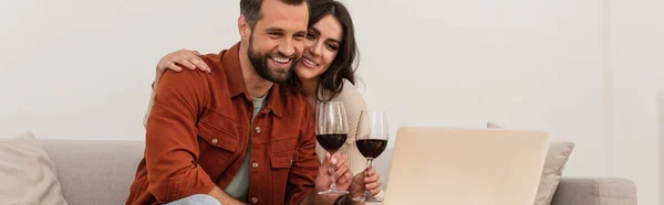 Mujer sonriente abrazando novio con copa de vino cerca de la computadora portátil, pancarta - foto de stock