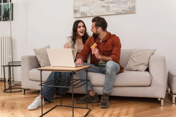 Femme excitée regardant petit ami avec carte de crédit près d'un ordinateur portable à la maison — Photo de stock