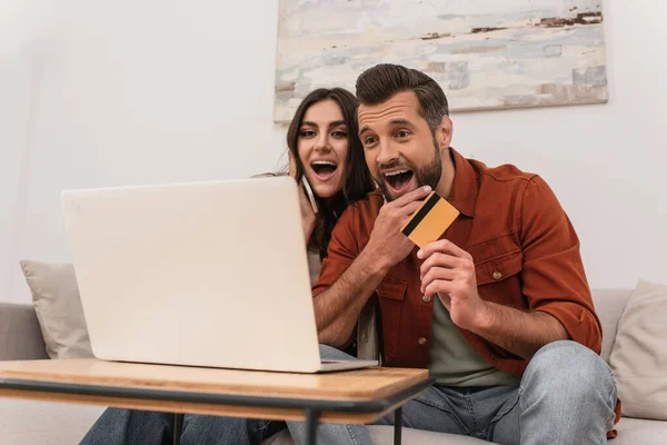 Восхитительная пара с кредитной картой и смартфоном смотрит на ноутбук дома — стоковое фото