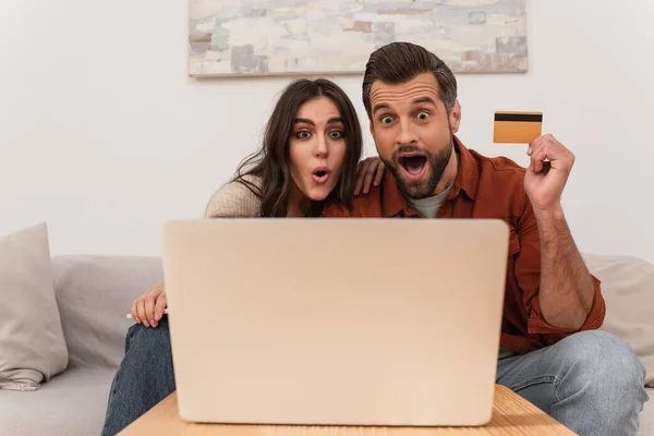 Casal surpreendido com cartão de crédito e smartphone olhando para laptop desfocado — Fotografia de Stock