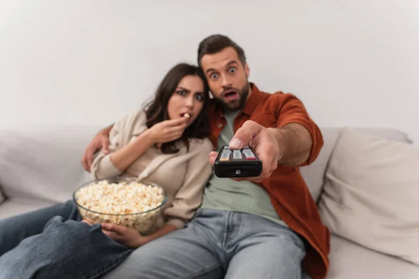 Fernbedienung in der Hand eines verschwommenen Mannes neben verblüffter Freundin mit Popcorn — Stockfoto