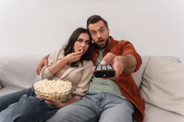 Télécommande en main de l'homme flou près de petite amie étonnée avec pop-corn — Photo de stock