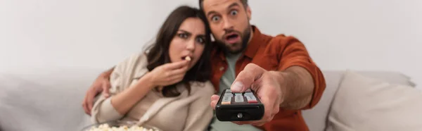 Mujer asombrada con palomitas de maíz sentado cerca de novio con controlador remoto en casa, pancarta - foto de stock