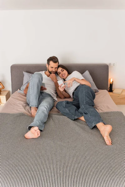 Улыбающаяся женщина, указывающая на смартфон, пока лежит рядом с парнем на кровати — стоковое фото