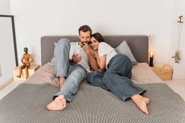 Smiling woman lying near boyfriend using smartphone on bed — Stock Photo