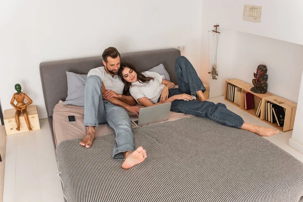 Smiling couple using laptop near smartphone on bed — Stock Photo