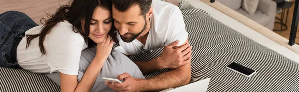 Couple using smartphone near blurred laptop on bed, banner — Stock Photo