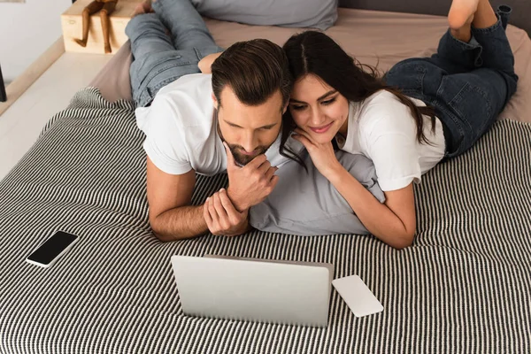Femme souriante regardant ordinateur portable près du petit ami et téléphone portable sur le lit — Photo de stock
