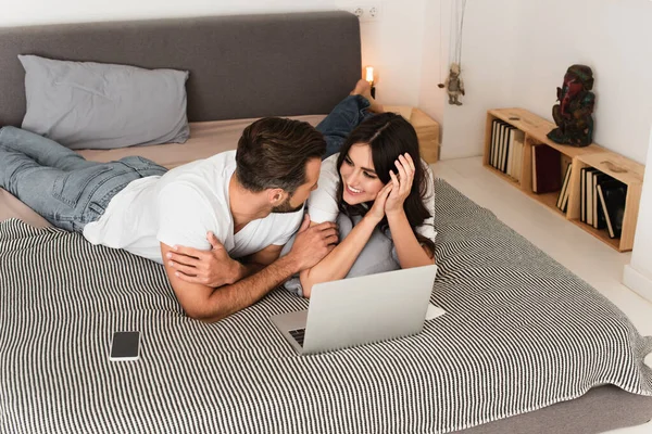 Couple joyeux regardant les uns les autres près des appareils sur le lit — Photo de stock