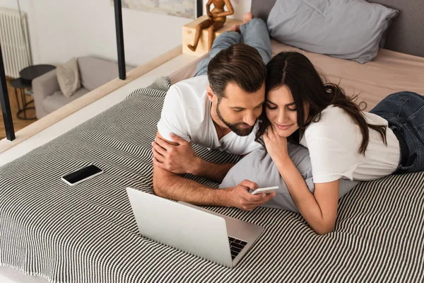 Homme utilisant téléphone mobile près de petite amie et ordinateur portable sur le lit — Photo de stock