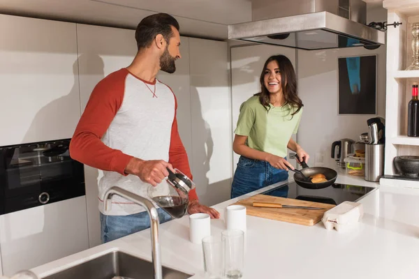 Улыбающаяся женщина готовит блинчики рядом с парнем с кофе на кухне — стоковое фото