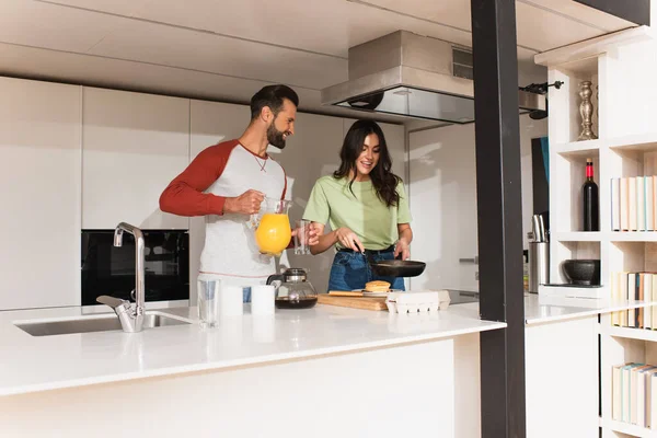 Sonriente hombre sosteniendo jarra con jugo de naranja cerca de novia cocinar tortitas - foto de stock