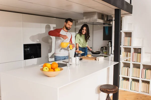 Man pouring orange juice near girlfriend with pancakes in kitchen — Stock Photo