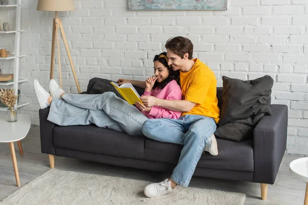 Счастливая молодая пара отдыхает на диване, обнимает и держит книгу в гостиной — стоковое фото