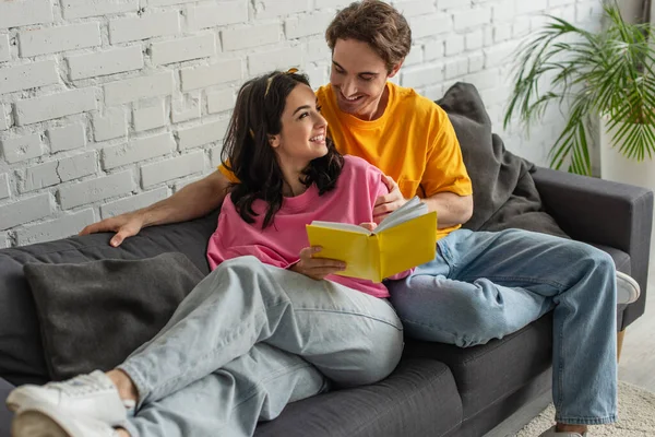 Sorrindo jovem casal descansando no sofá, abraçando e segurando livro na sala de estar — Fotografia de Stock