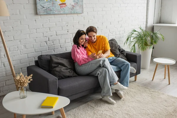 Lächelndes junges Paar sitzt auf Couch und schaut auf Handy im Wohnzimmer — Stockfoto