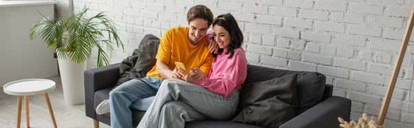 Smiling young couple sitting on couch and looking at cellphone in living room, banner — Stock Photo