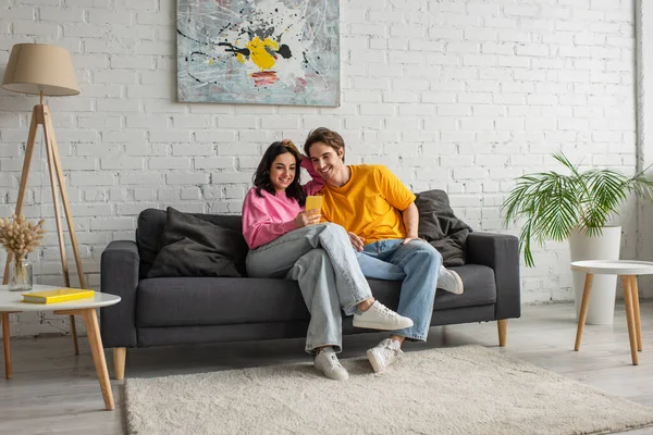 Sonriente joven pareja sentada en el sofá y mirando el teléfono celular en la sala de estar - foto de stock