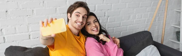 Sonriente joven pareja sentada en el sofá y tomando selfie con la lengua que sobresale en la sala de estar, bandera - foto de stock