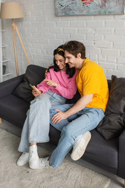 Sonriente mujer joven señalando con el dedo al teléfono celular cerca de novio en el sofá en la sala de estar - foto de stock
