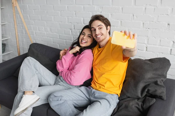 Smiling young couple sitting on couch and taking selfie with sticking out tongue in living room — Stock Photo