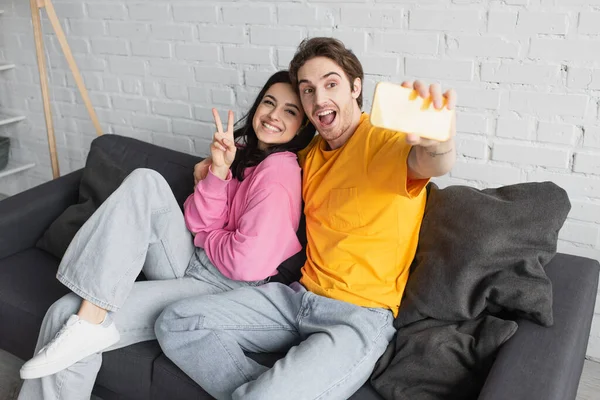 Sourire jeune couple assis sur le canapé et prendre selfie avec geste de paix dans le salon — Photo de stock