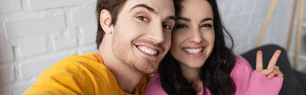 Sorrindo jovem casal sentado no sofá e olhando para a câmera com gesto de paz em casa, banner — Fotografia de Stock