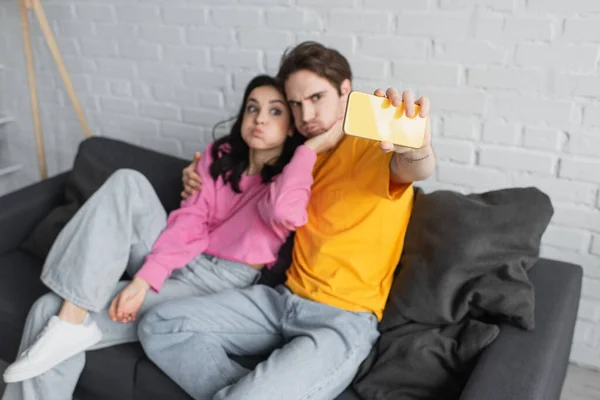 Sonriente joven pareja sentada en el sofá y tomando selfie y mueca en la sala de estar - foto de stock